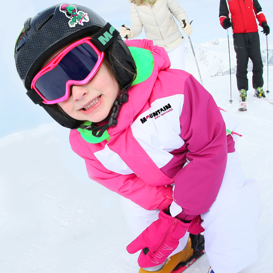 A smiling child, dressed in a helmet, goggles, and a cozy jacket from Mountain Kids Outfitters, is playing in the snow.