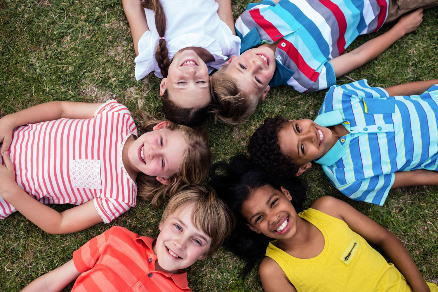 A group of children are lying on grass in a circle and smiling.