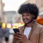 Young man standing on the street looking at his mobile phone and smiling